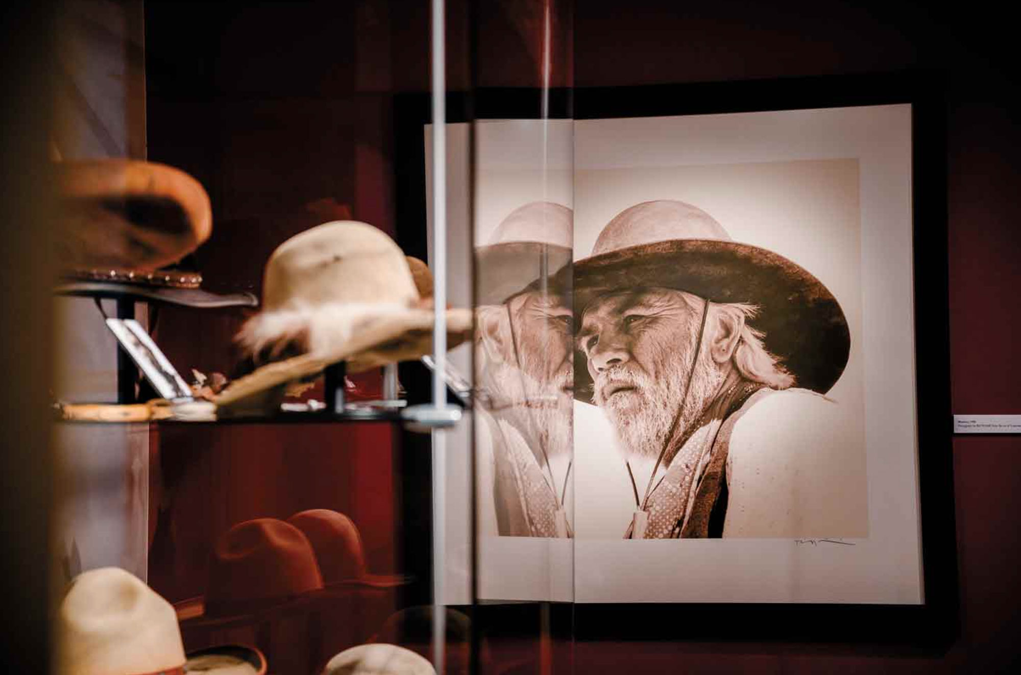 Hats sit stored in a clear display case. In the background is a blown up image hanging of an older man in a hat peering off into the distance